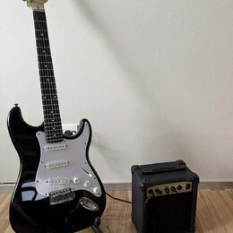 Černá elektrická kytara s bílým pickguardem a světle hnědým hmatníkem, umístěná na černém stojanu. Na hlavě kytary je viditelné logo Fender. Vedle ky…