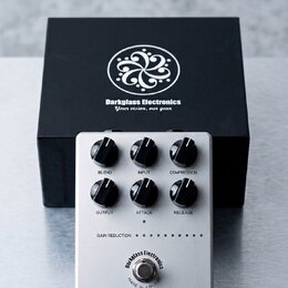 Stříbrný efektový pedál s šesti černými ovládacími knoflíky. Na spodní části je nápis SUPER SYMMETRY 115 GeV COMPRESSOR a kulaté logo Darkglass Elect…