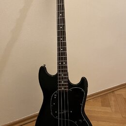 Černá elektrická basová kytara s hnědým hmatníkem a světlou dřevěnou hlavou. Na hlavě je nápis Fender MUSICMASTER BASS a logo Fender. Tělo má černý p…