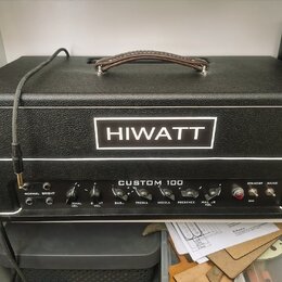 Černý kytarový zesilovač s koženou rukojetí a bílým lemováním. Na předním panelu je bílé logo HIWATT a nápis CUSTOM 100. Ovládací panel obsahuje přep…