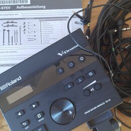 Na fotografii je černý elektronický bicí modul s nápisy Roland, V-Drums a DRUM SOUND MODULE TD-07. Vedle modulu jsou svázané černé kabely a černý nap…