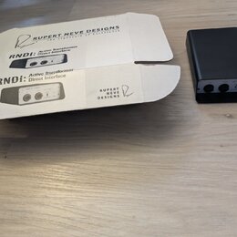 Na fotografii je černý, kompaktní box s dvěma vstupy a ovládacím prvkem. Vedle něj je otevřená kartonová krabice s nápisy RUPERT NEVE DESIGNS, The Si…