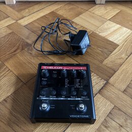 Černý efekt pedál s červeným pruhem a nápisy TC-HELICON harmony GXT a VOICETONE™. Zařízení obsahuje několik ovládacích knoflíků, přepínačů a displej…