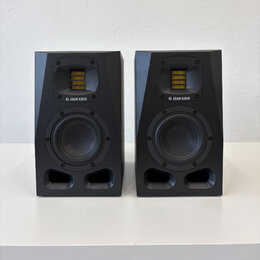 Dva černé studiové monitory značky ADAM AUDIO. Každý reproduktor má v horní části žlutý výškovek a ve spodní části černý basový reproduktor. Povrch j…