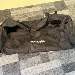 Černá transportní taška z textilního materiálu s několika úchopy. Na boku je bílý nápis ROCKBAG a pod ním menší text by Warwick. Taška je otevřená, p…