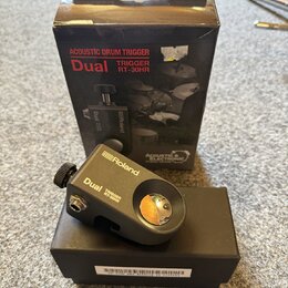 Černý akustický trigger na černém krabici a vedle originálního obalu. Na zařízení i obalu je bílé logo Roland a nápis Dual TRIGGER RT-30HR. Obal obsa…