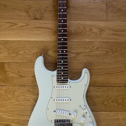 Bílá elektrická kytara s bílým pickguardem a hnedým hmatníkem. Na hlavě je viditelné logo Fender Stratocaster. Nástroj má tři snímače s bílými krytka…