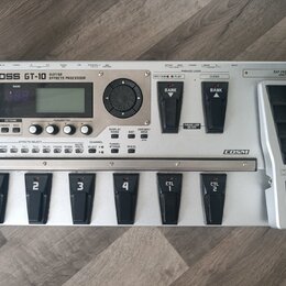 Stříbrný kytarový efektový procesor s nápisem BOSS GT-10 GUITAR EFFECTS PROCESSOR. Zařízení má digitální displej, několik ovládacích tlačítek a v pra…