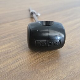 Černý plastový knoflík s nápisem Roland. Je připevněn na kovovou osa a položen na dřevěném povrchu. Povrch plastu vykazuje drobné škrábance.