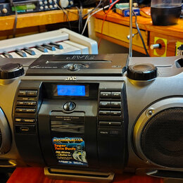 Šedý přenosný radiomagnetofon značky JVC. Na předním panelu je digitální displej, ovládací tlačítka a nápis POWERED BOOMBOX CD SYSTEM RV-KN401. Ve sp…