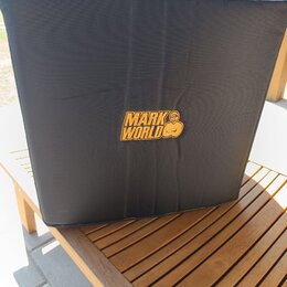 Černý textilní kryt na hudební zařízení, umístěný na dřevěném stole. Uprostřed je žluté logo s nápisem MARK WORLD a stylizovanou postavou. Povrch lát…