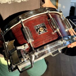 Červený dřevěný snare drum s kovovými obručemi a mechanismem na straně. Na korpusu je čitelný nápis TAMA SUPERSTAR a pod ním text MADE IN JAPAN. Povr…