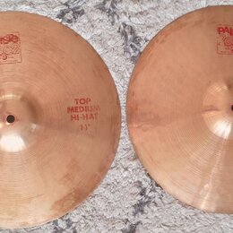Dva mosazné činely typu hi-hat položené na šedém koberci. Levý činel nese nápisy „PAISTE 2002“, „TRADE MARK MADE IN SWITZERLAND“ a „TOP MEDIUM HI-HAT…