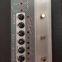 Stříbrný kytarový pedál s kovovým šasi a texturovaným horním panelem. Na přístroji jsou nápisy TECH 21 NYC, SANSAMP a VT Bass DELUXE. Obsahuje šest č…