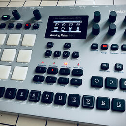 Stříbrný digitální drum machine s názvem Analog Rytm MKII a logem Elektron. Zařízení obsahuje displej, zahlreiche ovládací knofíky, bílé gumové pady…
