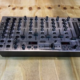 Stříbrný DJ mixér s černými ovladači a posuvníky. Na levém boku je nápis VMX 1000 a na pravém boku logo BEHRINGER. Uprostřed spodní části je nápis XP…