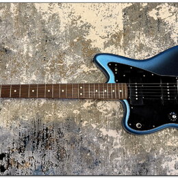 Modrá elektrická kytara s černým pickguardem a hnědým hmatníkem. Na hlavě je viditelné logo Fender a nápis Stratocaster. Kytara má jeden humbucker u…