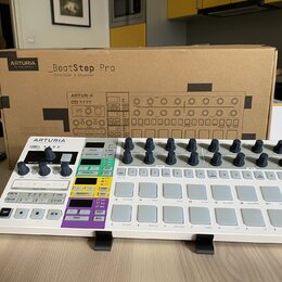 Bílý sekvencer Arturia BeatStep Pro umístěný na černém stojanu před svou originální kartonovou krabicí. Na přístroji jsou vidět šeradovky šedých pods…