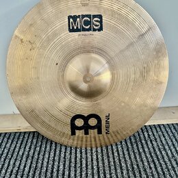 Zlatavý kovový činel s viditelným broušením. Na povrchu jsou nápisy MCS a 20" Medium Ride, v dolní části je logo MEINL. V horní části je malý nečitel…