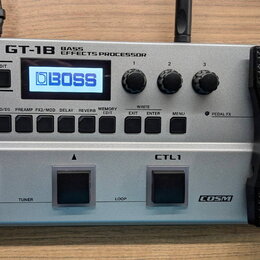 Stříbrný basový efektový procesor s černým výrazným pedálem vpravo. Na horní části je nápis BOSS GT-1B BASS EFFECTS PROCESSOR a na displeji logo BOSS…