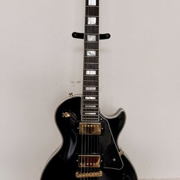 Černá elektrická kytara s gold hardwarem umístěná na černém stojanu. Na hlavě hmatníku je bílý nápis Epiphone. Kytara má dva humbuckery, zlatý most a…