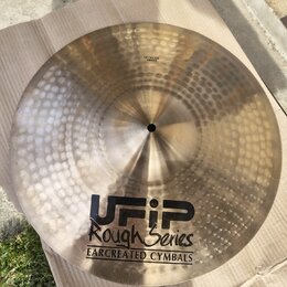 Zlatavý kovový činel s kladivkovaným povrchem, položený na hnědém kartonu. Na povrchu je černý nápis UFIP Rough Series EARCREATED CYMBALS a malá nále…