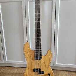 Pětstrunná basová kytara s přírodním dřevěným povrchem těla a hmatníkem z tmavého dřeva. Na hlavě nástroje je svisle napsáno „Marcus Miller Deluxe 5“…
