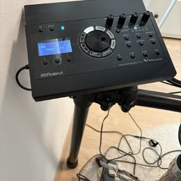 Černý elektronický bicí modul s displejem, na kterém je nápis "KIT 100 User Kit". Na spodní hraně je logo "Roland" a vpravo nápis "DRUM SOUND MODULE…