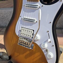 Elektrická kytara s tělem v provedení sunburst a bílým pickguardem. Nástroj má tři snímače typu single-coil, chromovaný most s páčkou a tři bílé ovlá…