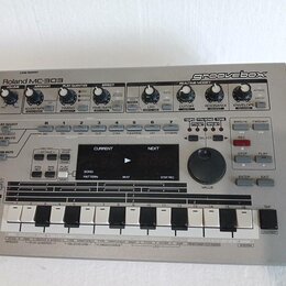 Stříbrný sekvenční syntezátor Roland MC-303 groovebox. Na předním panelu jsou umístěny ovládací knofíky, tlačítka a displej s nápisy jako CURRENT, NE…