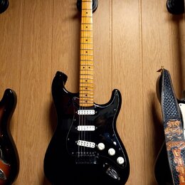 Černá elektrická kytara s hmatníkem ze světlého dřeva. Na hlavě je viditelné logo Fender. Kytara má tři snímače (dvě humbuckery a jeden jednosnímač),…