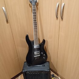 Černá elektrická kytara s dvojstupňovými humbuckery, opřená o černý kytarový kombo zesilovač. Na hlavě kytary je bílý nápis Cort. Zesilovač má na pře…