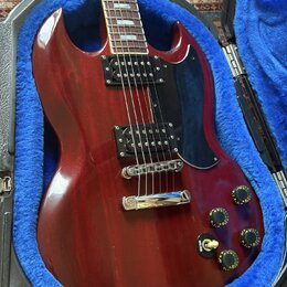 Kytara s červeným lakem a černým plechtrátem, umístěná v otevřeném kufru s modrou výplní. Nástroj má dva černé humbuckery, chromovaný most a čtyři če…