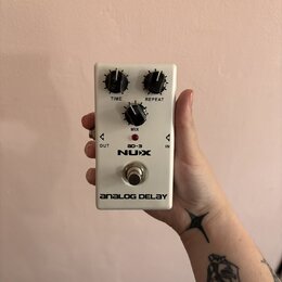 Bílý kytarový efekt s nápisy NUX, ad-3 a ANALOG DELAY. Na horní straně jsou tři černé otočné ovnovky s popisky TIME, REPEAT a MIX. V dolní části je k…