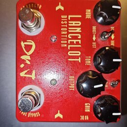 Červený kytarový efekt s nápisy LANCELOT DISTORTION a D.T. na horní straně jsou čtyři černé otočné knoflíky s popisky MORE, TONE, OUTPUT a GAIN. Na p…