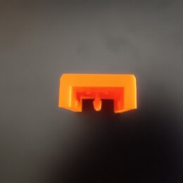 Na fotografii je malý, oranžový plastový dílek obdélníkového tvaru s výřezem v dolní části. Povrch je hladký a bez viditelných nápisů či log. Dílek v…