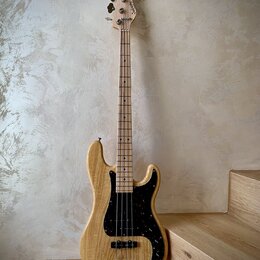 Elektrická basová kytara s přírodním dřevěným povrchem těla a hmatníku. Nástroj má černý pickguard s bílými tečkami, dva snímače a čtyři ladičky. Na…