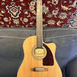 Akustická kytara s vyřezem v těle, přírodní dřevěnou barvou a hnědým hmatníkem. Na hlavě je bílý nápis Epiphone a na štítku uvnitř otvoru zvukové dír…