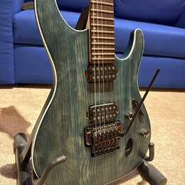 Elektrická kytara s modrozeleným průsvitným povrchem, pod kterým je vidět výrazná struktura dřeva. Nástroj má bílý lem kolem korpusu, dva humbuckery…