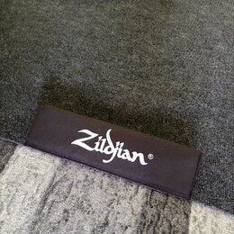 Fotografie zachicuje detail černé látkové plochy, pravděpodobně koberce nebo podložky. Na ní je bílým tiskem uveden nápis Zildjian®. Povrch materiálu…