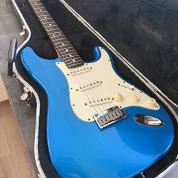 Modrá elektrická kytara typu Stratocaster s krémovým pickguardem a hnedým hmatníkem, umístěná v otevřeném tvrdém kufru. Na hlavě krku je viditelné lo…