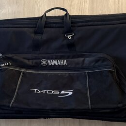 Černý polstrovaný pouzdro na klávesový nástroj s rukojetí a popruhy. Na přední části je připevněna černá taška s nápisy YAMAHA a Tyros 5. Povrch pouz…