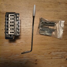 Kytarový kobylník stříbrné barvy s nápisem Floyd Rose. Vedle něj je umístěna páka s bílým koncem a plastový sáček s montážním příslušenstvím, včetně…