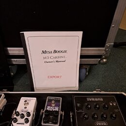 Na fotografii je otevřený transportní kufr s několika efektovými pedály a uživatelskou příručkou. Na bílém sešitě je nápis MESA/BOOGIE, M3 CARBINE. O…