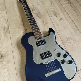 Tmavě modrá elektrická kytara s kovovým pickguardem a hnědým hmatníkem. Nástroj má dva humbuckery, tři ovládací knofíky a jeden přepínač. Na hlavě kr…