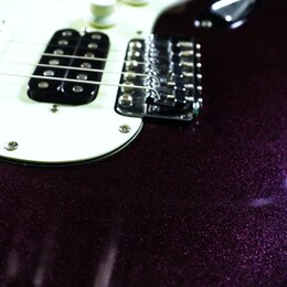 Detail elektrické kytary s tmavě fialovým metalizovaným povrchem s třpytivým efektem. Nástroj má bílý pickguard, tři snímače (dva jednoduché a jeden…