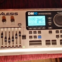 Stříbrný elektronický bicí modul značky ALESIS, model DM10. Na přední straně je displej s nápisem Fusion a Tempo 24 BPM, pět posuvníků pro ovládání h…
