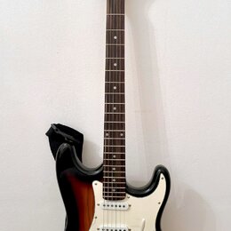 Elektrická kytara s tłem v provedení sunburst a bílým pickguardem. Na hlavě hředu je viditelné logo "Squier". Nástroj má tři snímače typu single-coil…