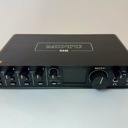 Černý audio rozhraní s nápisem MOTU M6 na horní straně. Přední panel obsahuje čtyři ovratky GAIN, tlačítka MON a jeden velký ovládací knoflík s nápis…