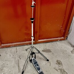 Stříbrný kovový stativ s černými plastovými prvky a rozkládacím trojnohým základem. Na stativu je umístěna černá efektová pedálka s bílým logem ve tv…